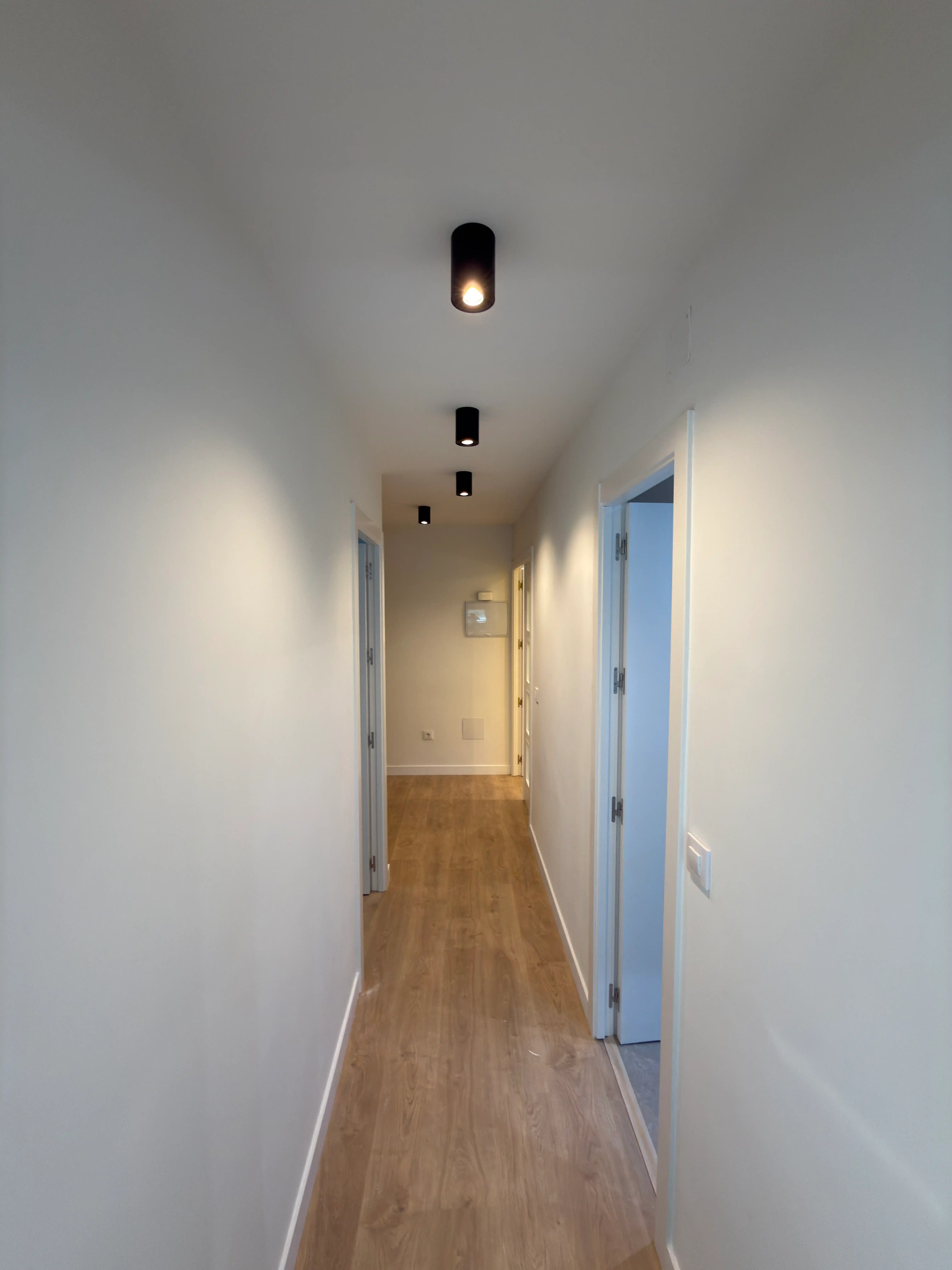 Pasillo interior con suelos de madera, puertas azules e iluminación LED de superficie.
