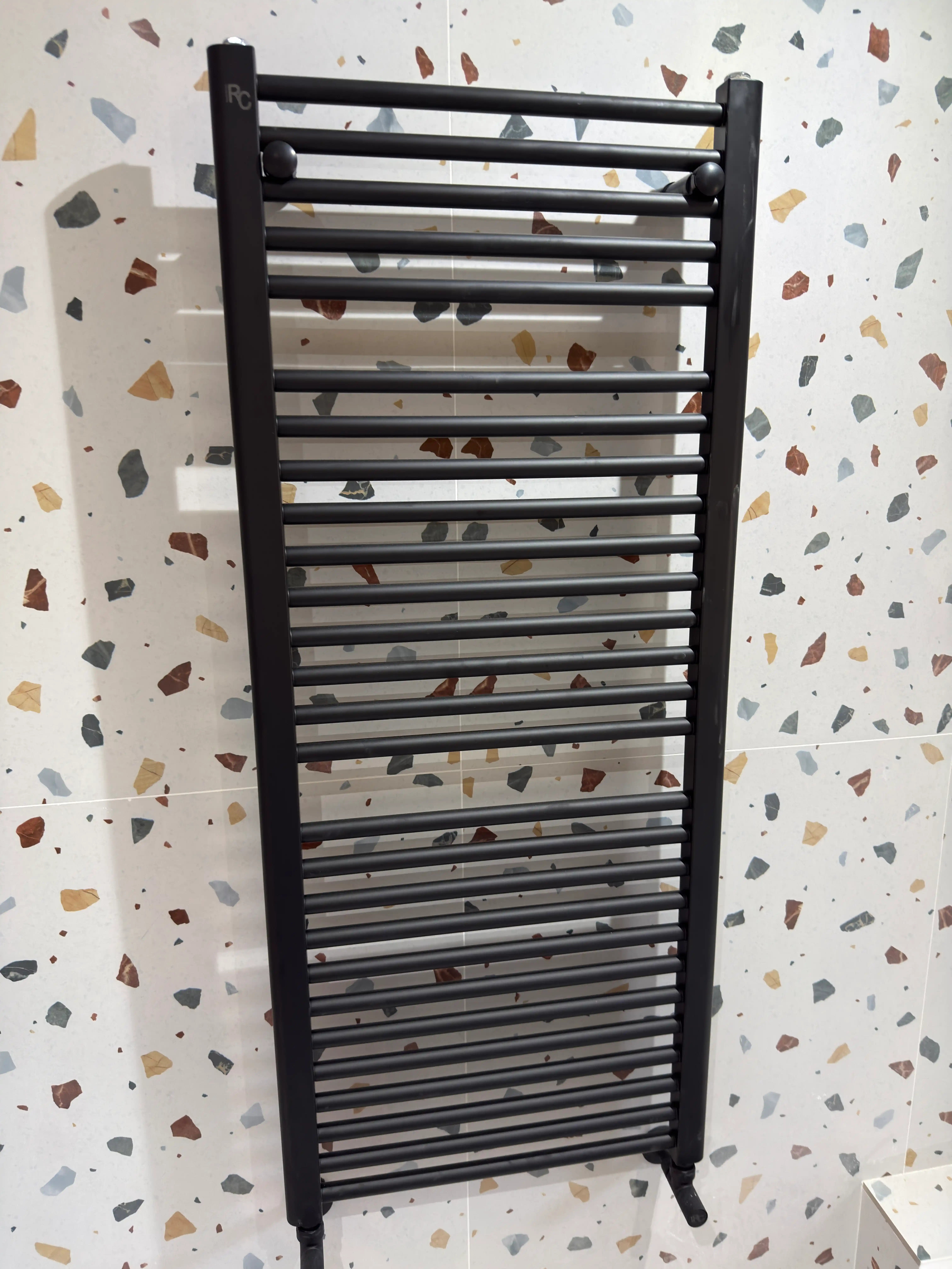 Radiador toallero negro mate instalado sobre pared con revestimiento cerámico estilo terrazo.